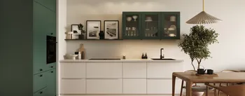 Moderne Küchenzeile in Grün und Weiß mit Glas-Hängeschränken, offener Ablage mit gerahmten Fotos, schwarzem Spülbecken und Armatur; links grüner Hochschrank mit Backofen, rechts Holztisch und Zimmerbaum, Fischgrät-Parkett und Deckenstrahler.